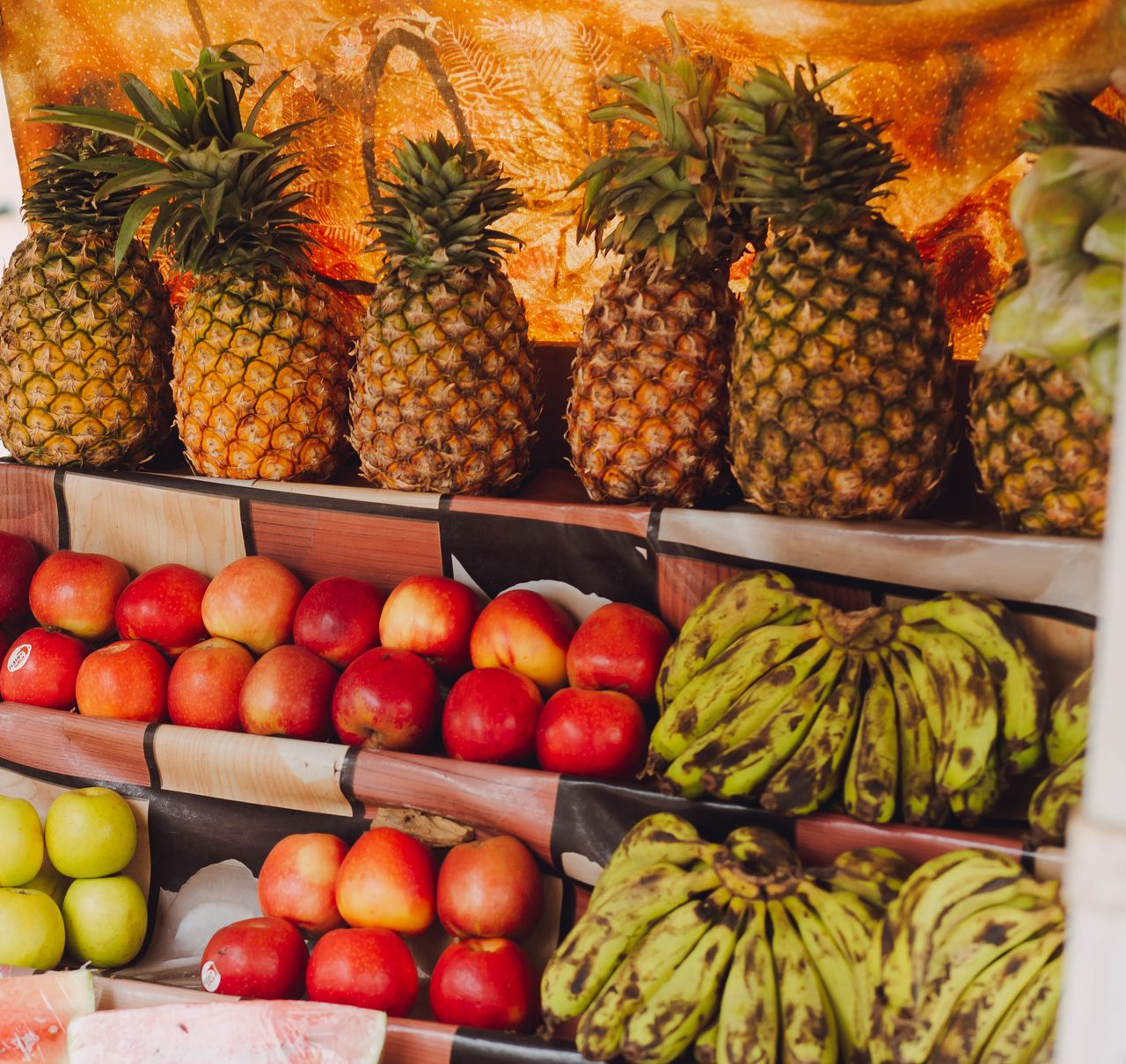 A colorful display of pineapples, apples, and plantains at a fruit market stand.